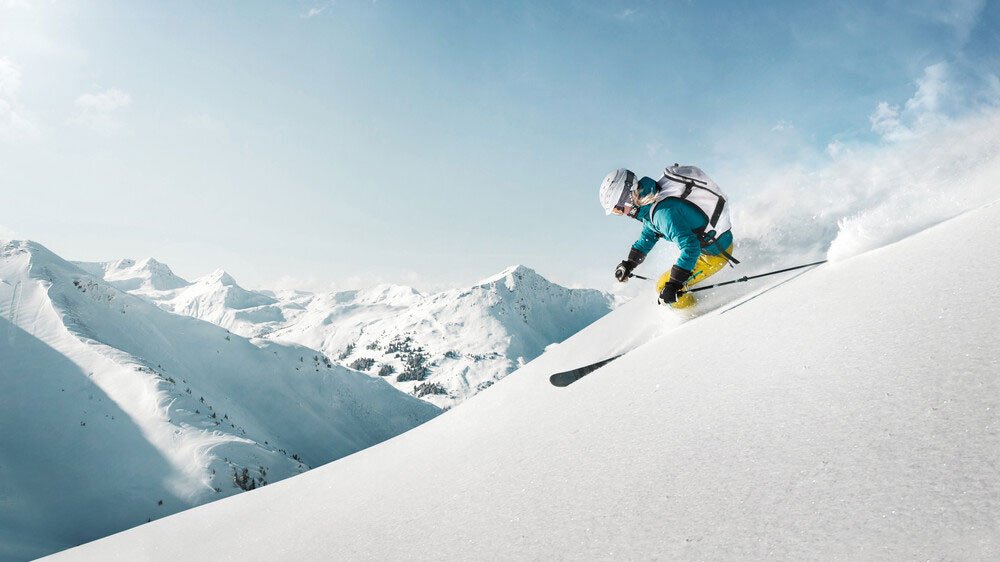 A skier dressed in blue and yellow gear skiing down a snowy mountain slope with snow-covered peaks in the background.