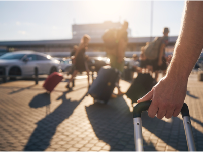 Person holding a rolling suitcase at an airport, with travelers and luggage in the background and sunlight casting shadows.