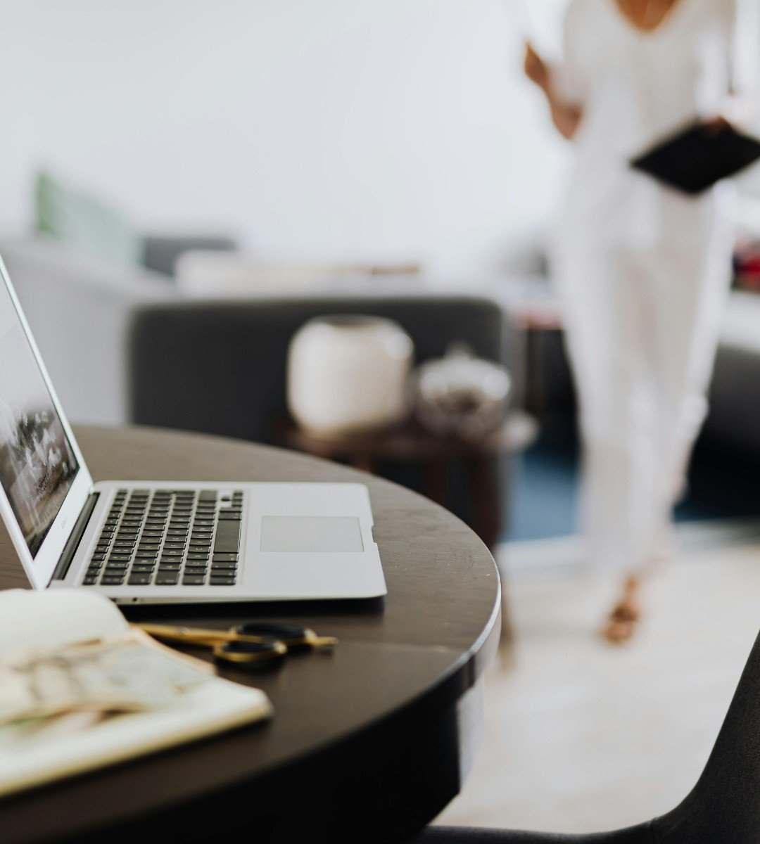 Open laptop on a round wooden table with a notepad, pen, and scissors in the foreground, blurred woman holding a tablet in the background.