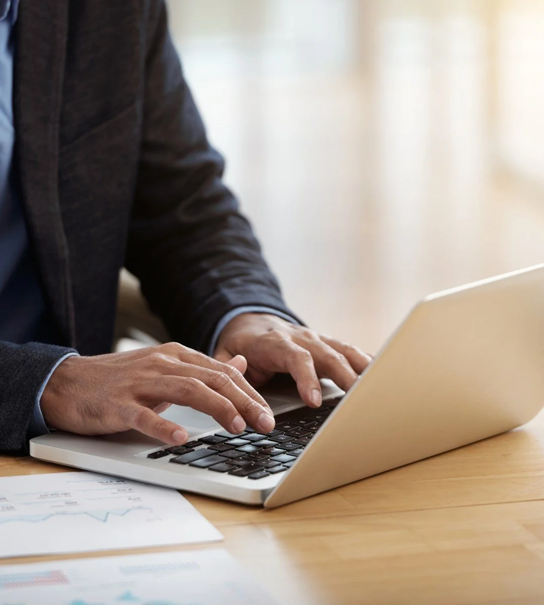 Person in a suit typing on a silver laptop at a wooden desk.
