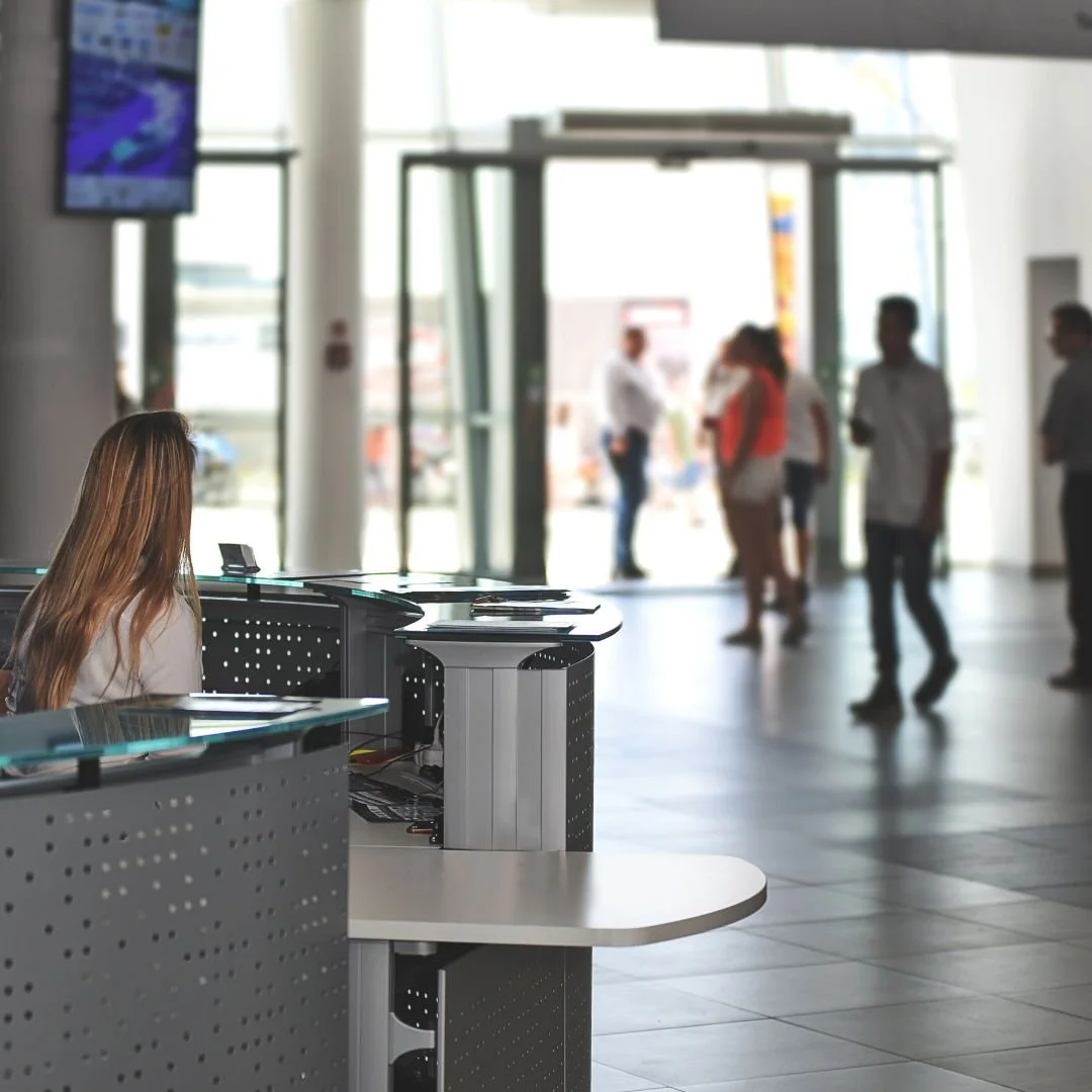 A woman sitting at a desk inside a modern airport or transportation hub, with a glass entrance in the background and people walking outside.