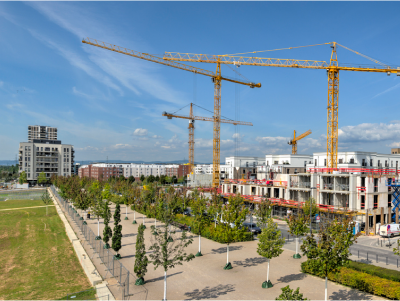 Construction site with several tall yellow cranes and partially built structures, alongside a park with trees and walkways, in an urban area under a clear blue sky.