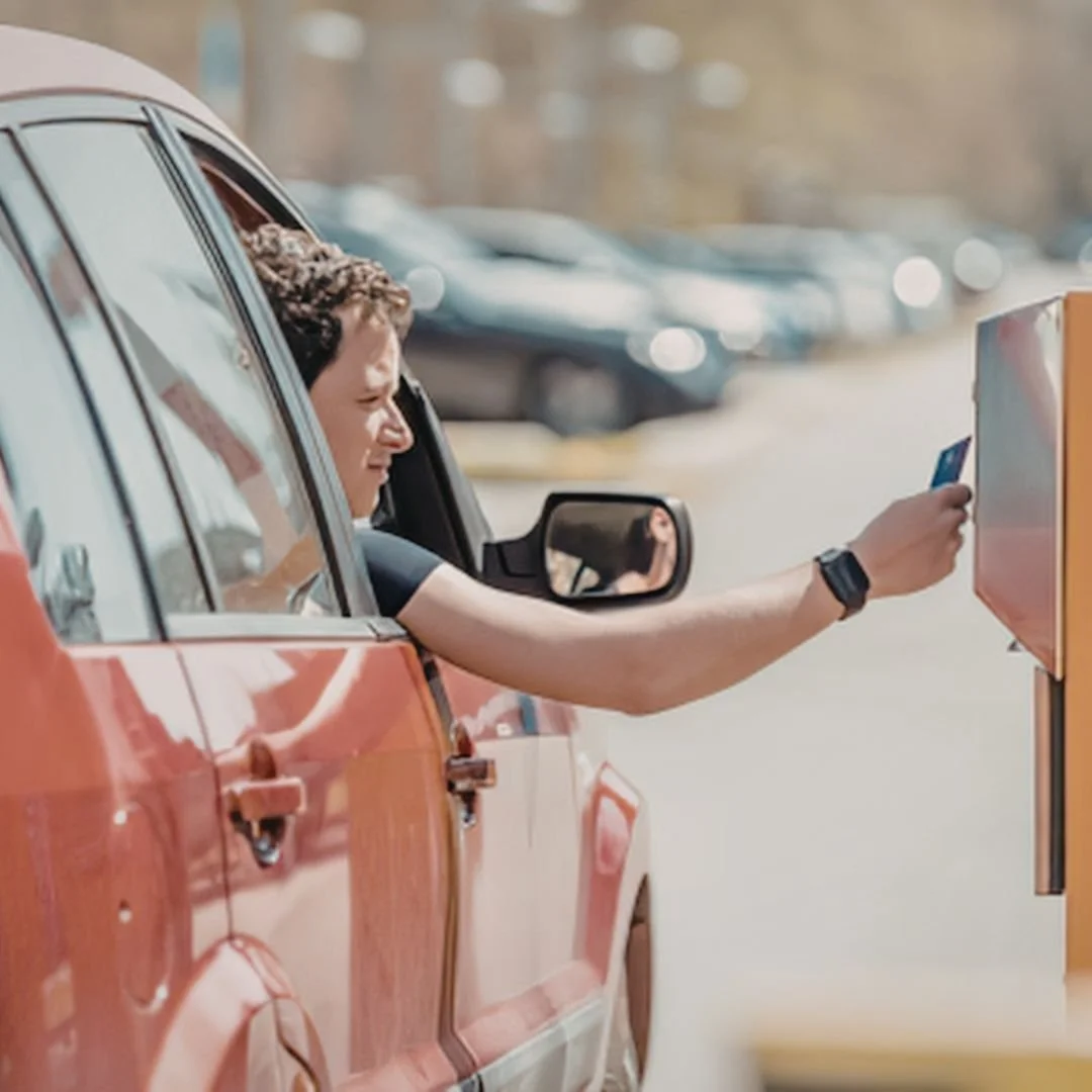 A person in a red vintage car inserting a credit card into a payment kiosk outside.