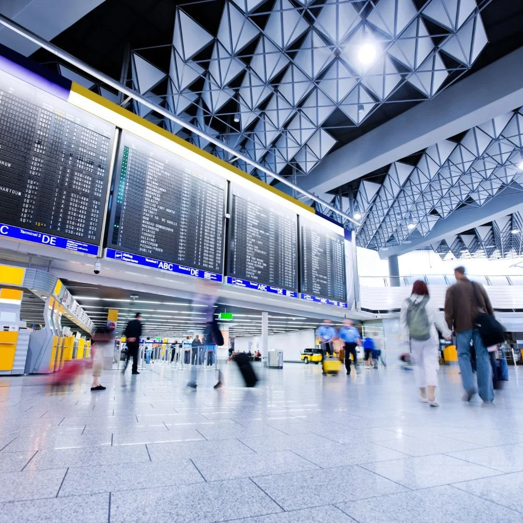 An airport terminal with large departure boards, travelers walking with luggage, and modern architectural ceiling design.