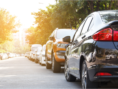 Line of parked cars along a street with trees and sunlight in the background.