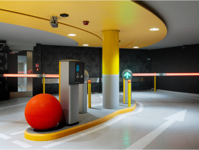 Entrance to a parking garage with a yellow ceiling, white and yellow curb, a red ball, a parking payment kiosk, and a green arrow sign indicating direction.