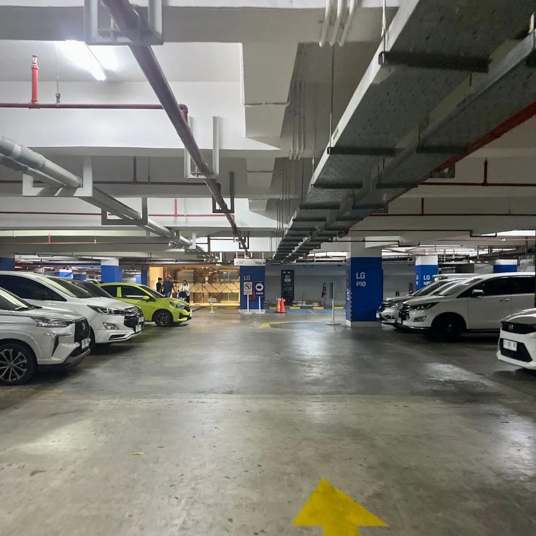 Underground parking garage with several parked cars, some are white, one is yellow, and one is black. Parking signs indicate sections LG P10 and LG N7, with overhead pipes and electrical conduits visible.