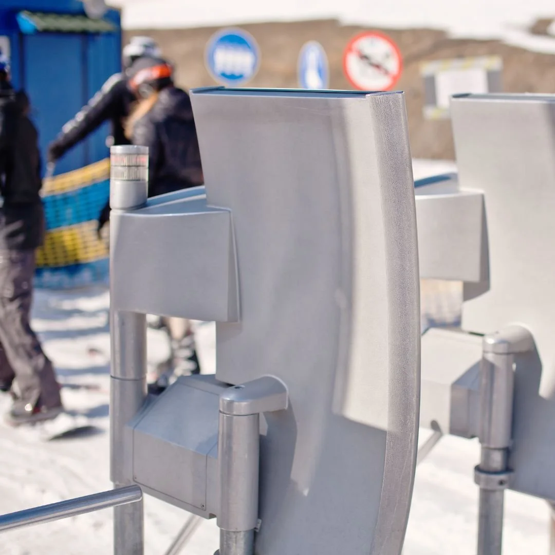 Close-up of a metal turnstile at a snowy outdoor location, with people in winter clothing in the background and signs visible on a hill.