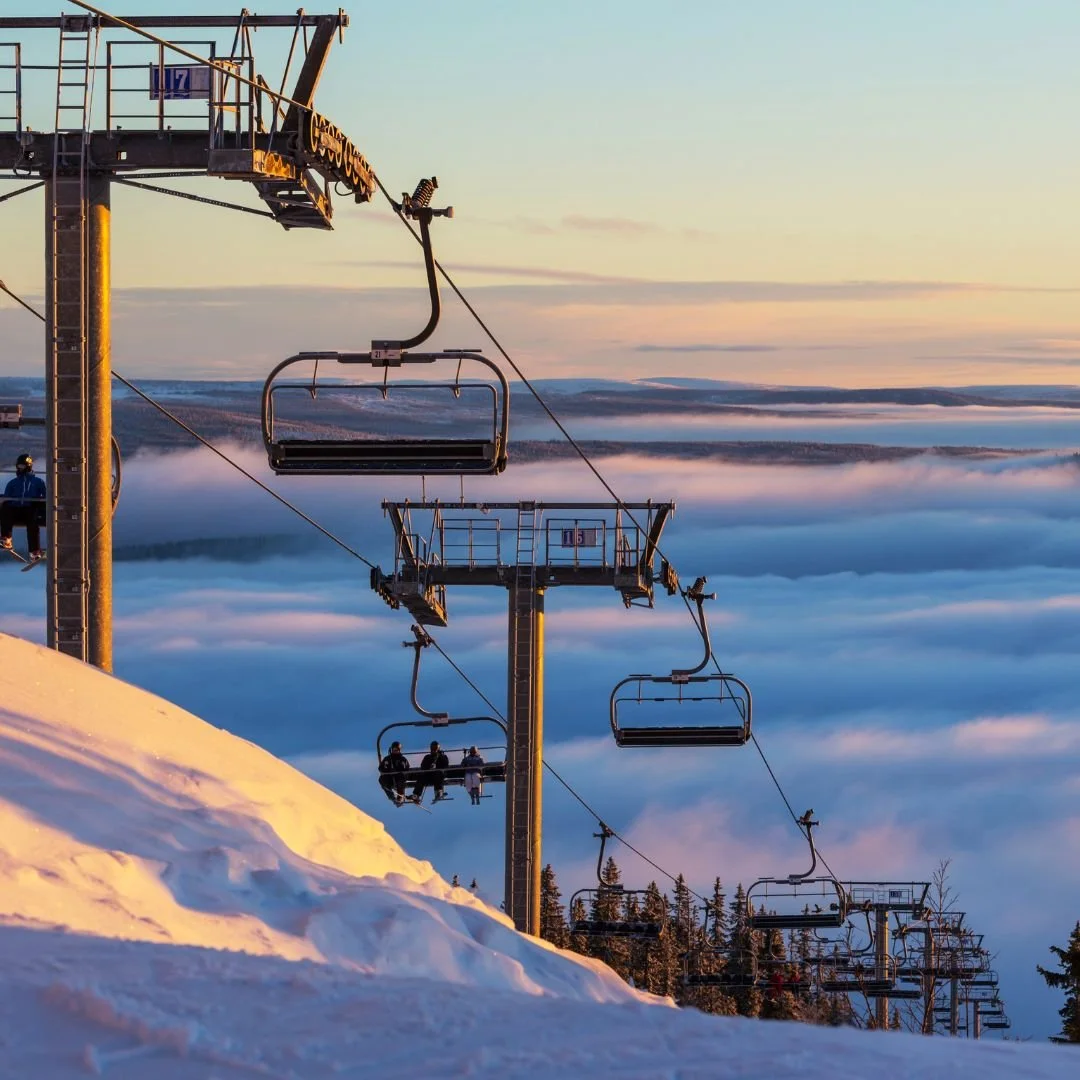 Ski lift chairs ascending over snow-covered slopes with a scenic view of clouds and distant mountains at sunrise or sunset.