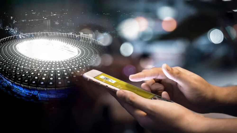 Person using a smartphone with a digital concert or event ticket displayed, with a brightly lit stage or arena in the background, possibly in a vehicle or at night.