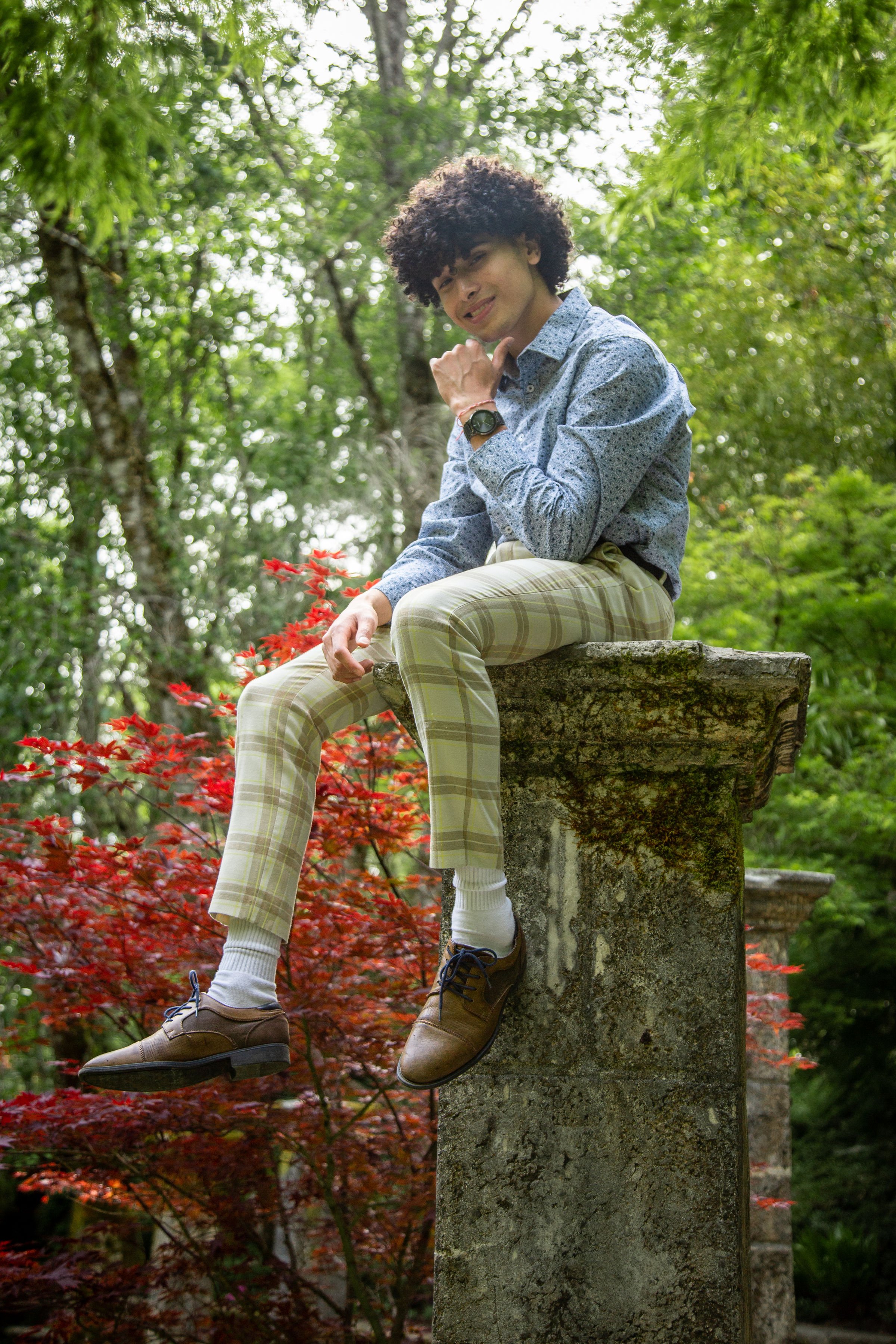 A young man with curly hair wearing a blue patterned shirt and plaid beige pants sitting on a stone ledge in a lush green outdoor setting with red foliage.