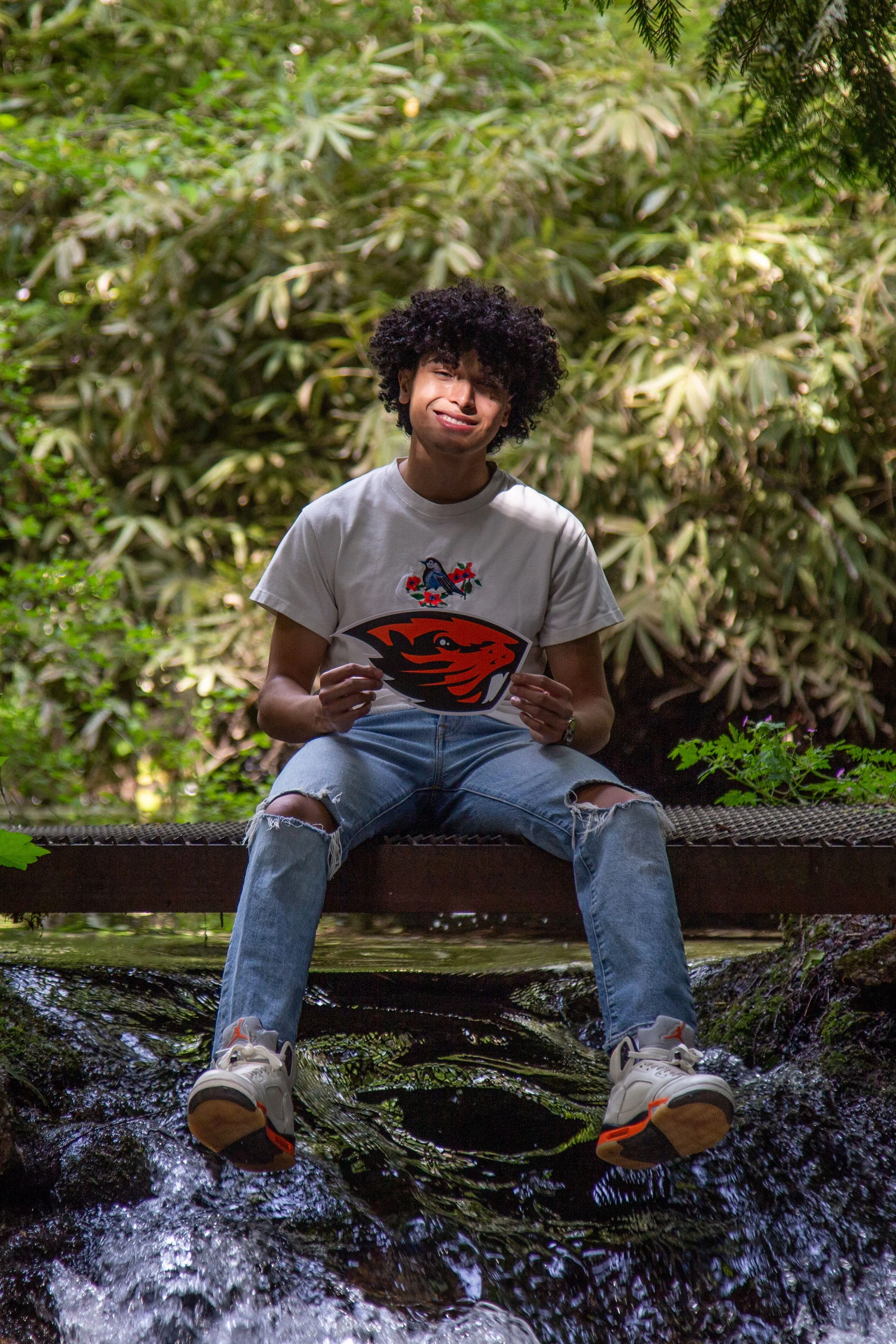 Person sitting on a wooden railing over a small stream, holding a sticker of a lion's head logo, surrounded by lush green foliage.