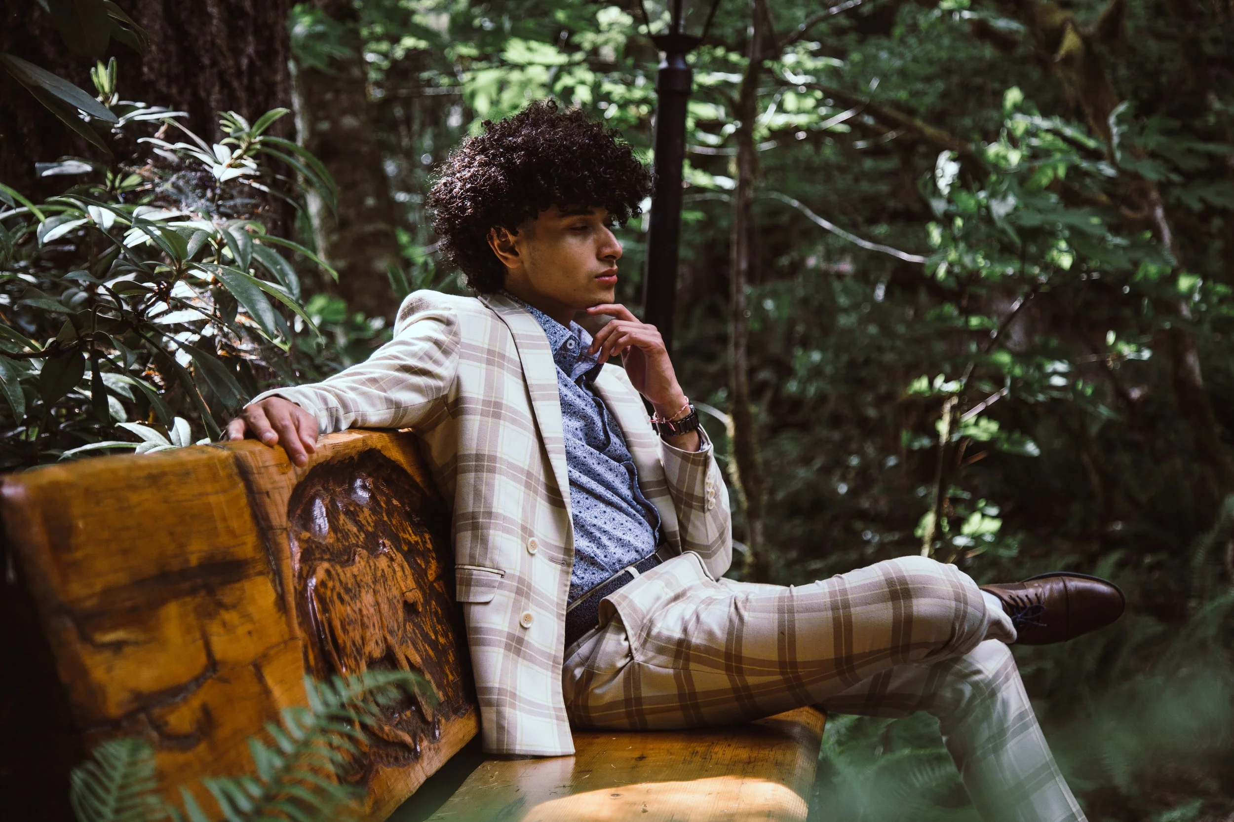 A young man in a beige plaid suit sitting on a wooden bench in a dense forest, with his hand on his chin, looking thoughtful.