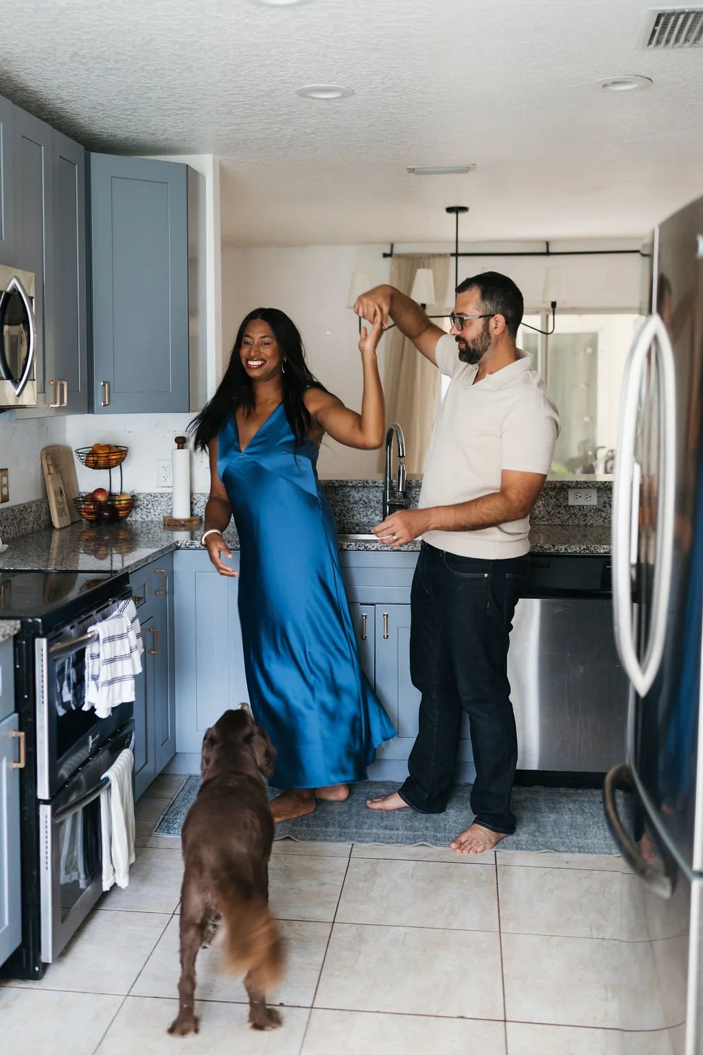 A man and woman are dancing together in a kitchen, with the woman smiling and wearing a blue dress, and the man with glasses wearing a white shirt and black pants. A brown dog watches them from the floor.