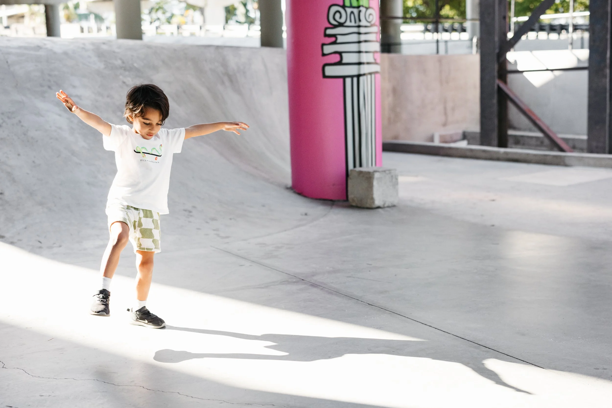A young boy with dark hair, wearing a white t-shirt, patterned shorts, and sneakers, balancing on a skateboard at a skate park.