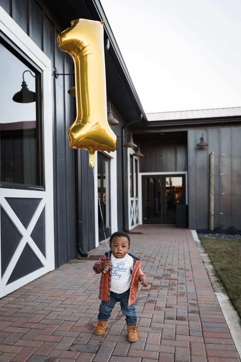 A young boy standing outdoors on a brick pathway, holding a large, gold, helium balloon shaped like the number one, in front of a dark building with white-framed windows and doors, during daytime.