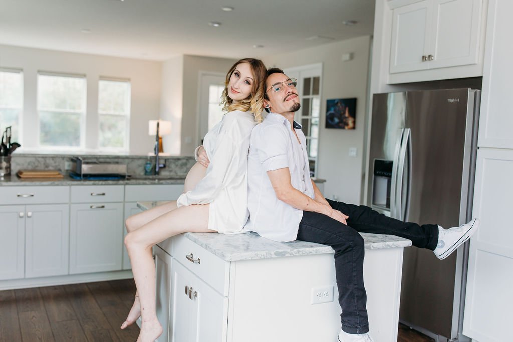 A woman and a man pose playfully in a bright kitchen, with the woman sitting on the kitchen counter and the man sitting on the island, both smiling.