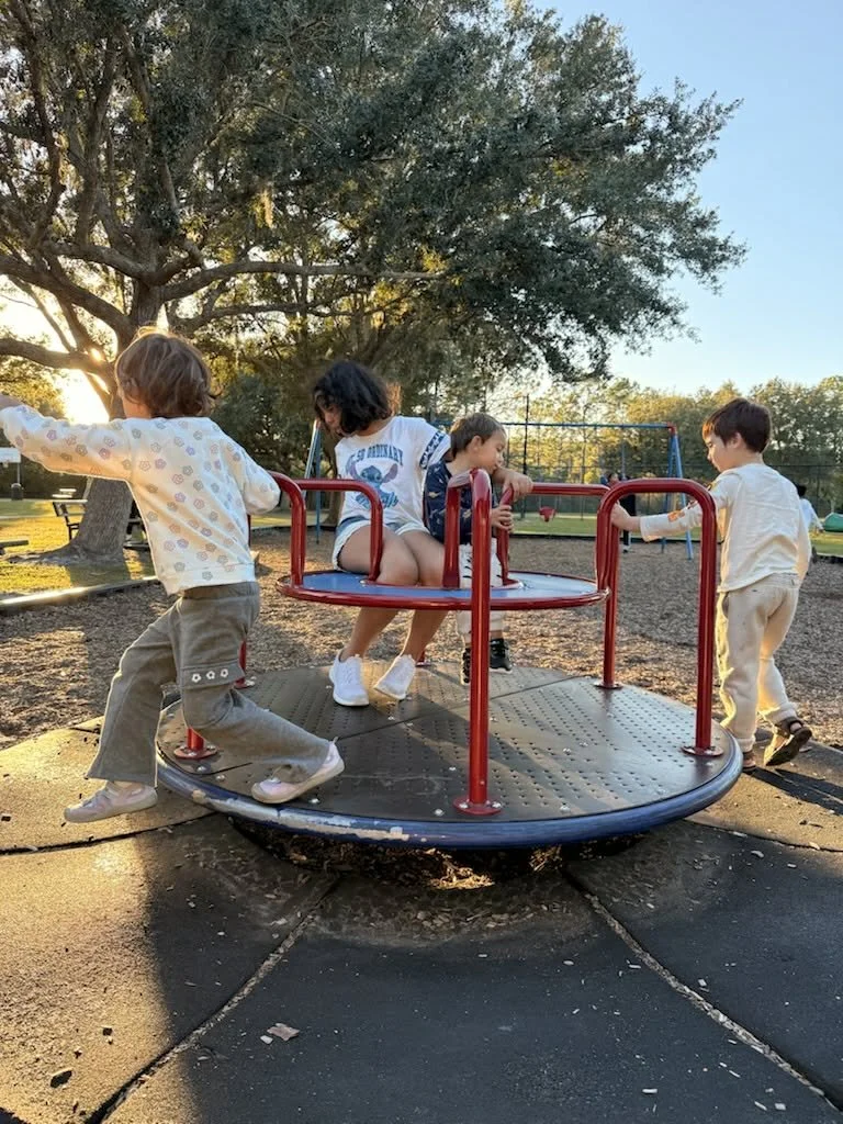 Four children playing on a merry-go-round at a park during sunset with trees and a clear sky in the background.