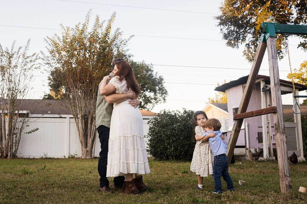 A woman and a man hugging in a backyard, two children, a girl and a boy, hugging near a swing set, with trees and a house in the background during sunset.