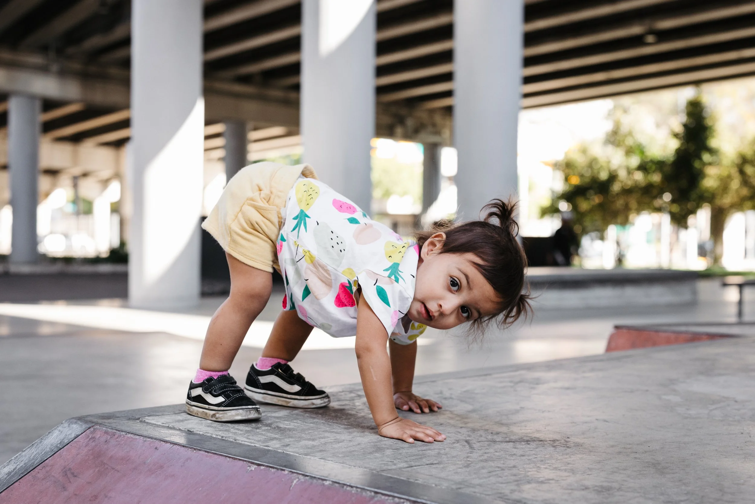 Young girl in colorful shirt and shorts crawling on a skatepark ramp under a highway overpass