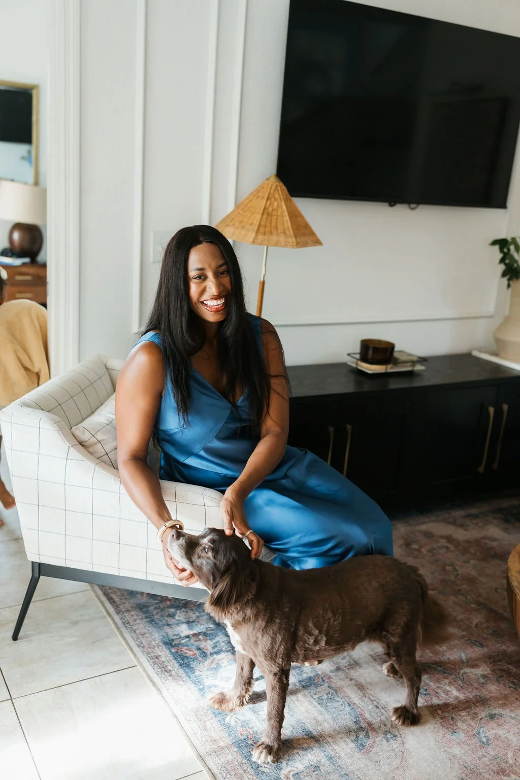 A woman in a blue dress sitting on a checkered armchair, smiling while petting a brown and white puppy in a living room with a large flat-screen TV, a lamp, and decorative items.