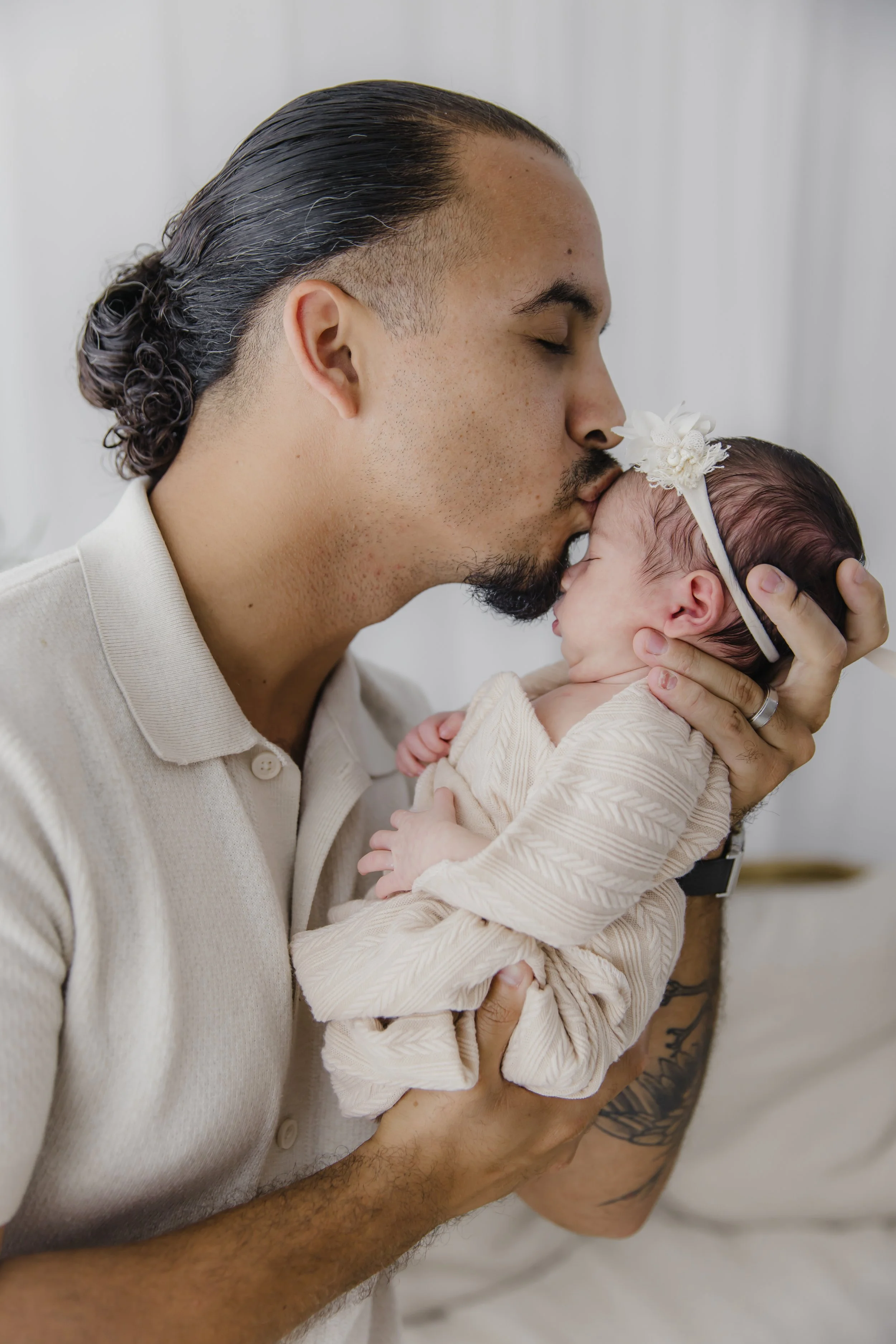 A man with dark hair tied back and a beard gently kisses a newborn baby on the forehead, holding her close. The baby wears a cream-colored outfit and a headband with a flower.