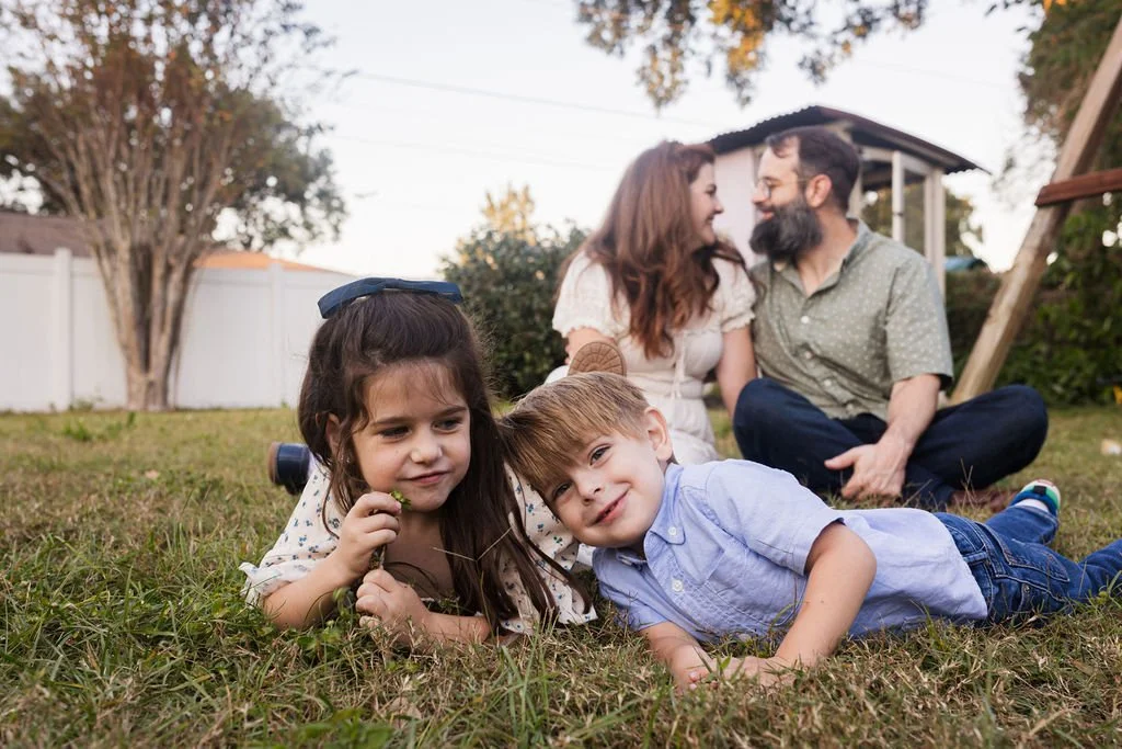 A family of four enjoying time outdoors in a backyard, with two children lying on the grass in the foreground and a woman and man sitting behind them, smiling and looking at each other.