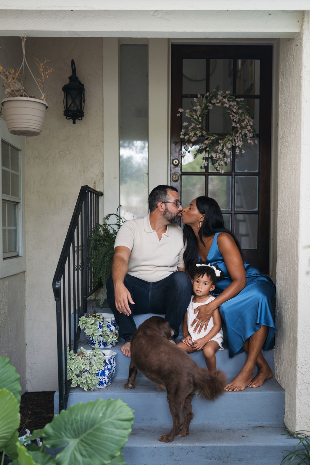 A couple sharing a kiss on their front porch with their young daughter and dog, decorated with a floral wreath on the door and potted plants nearby.
