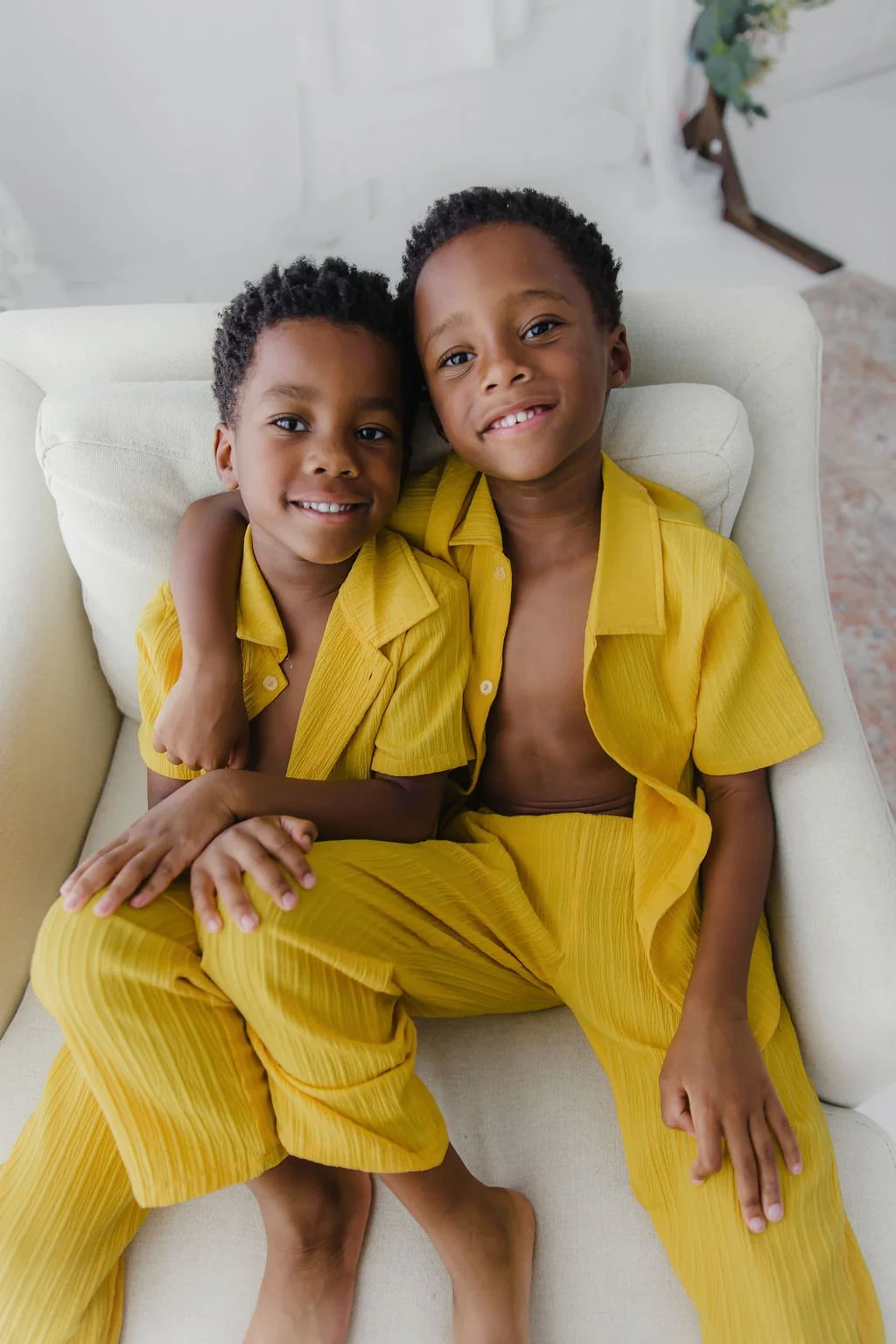 Two young boys sitting on a cream-colored sofa, smiling, with one boy's arm around the other. They are both wearing yellow matching pajamas.