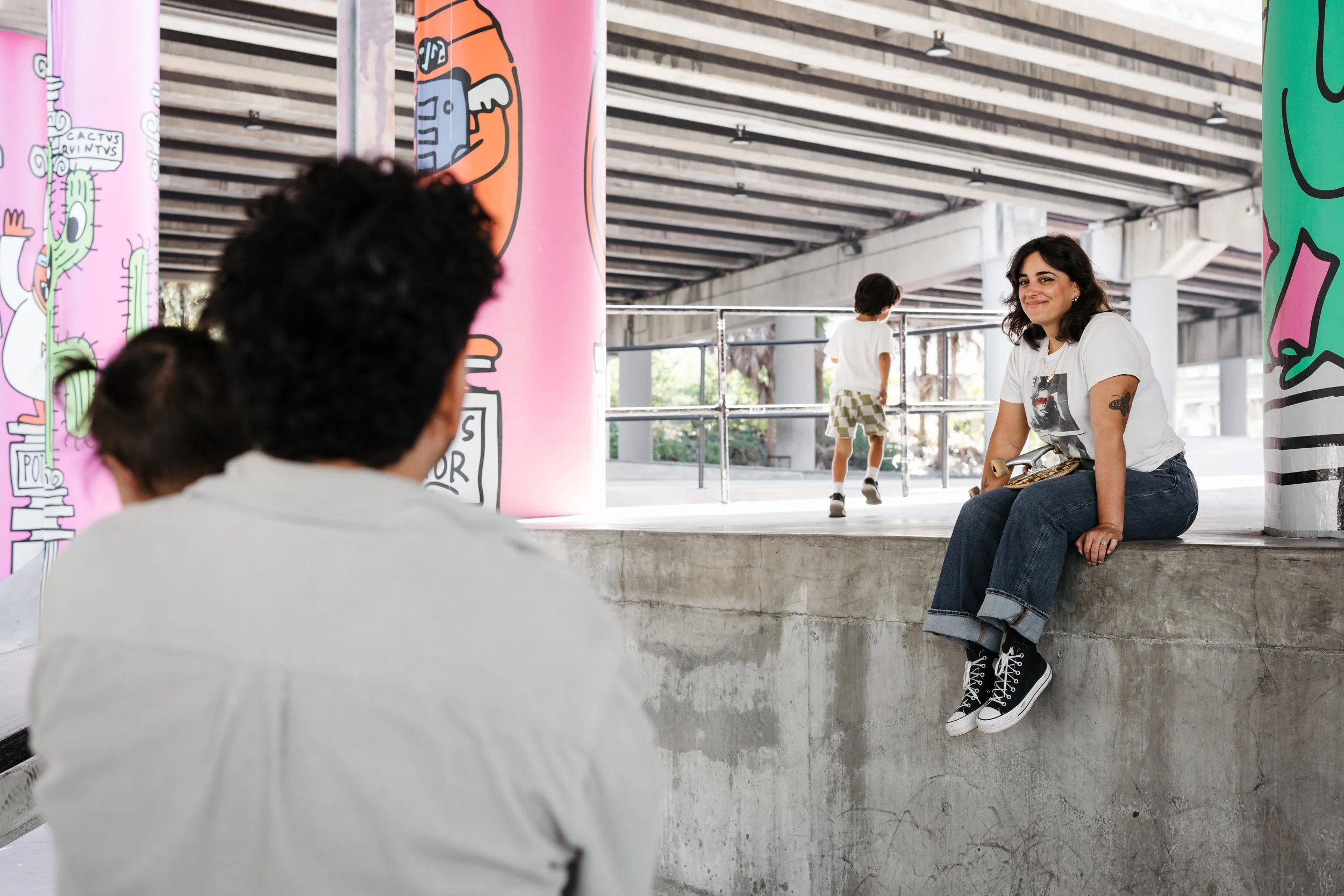 A woman sitting on a concrete ledge with skateboard, smiling, under a bridge decorated with colorful graffiti, with two children playing in the background.