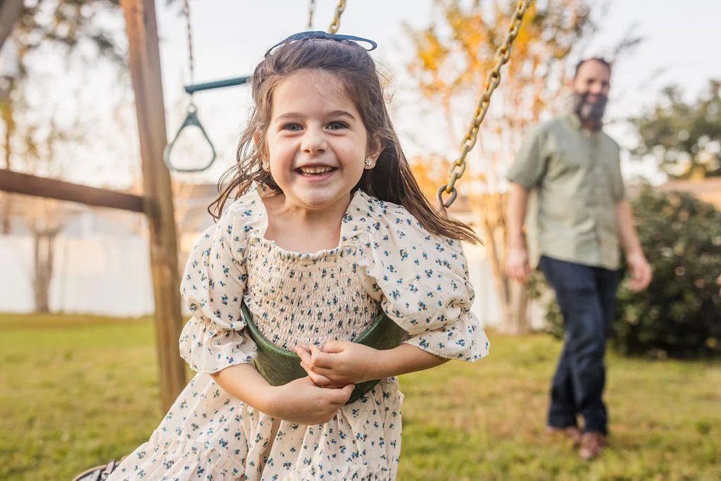 A young girl in a floral dress smiling in a park playground, with an adult man in the background near trees and greenery.