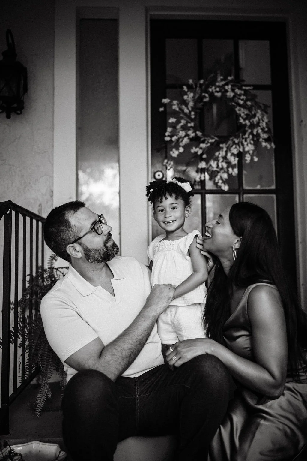 A happy family of three, a man, woman, and young girl, sitting together outside near a door decorated with a wreath, smiling and enjoying each other's company.