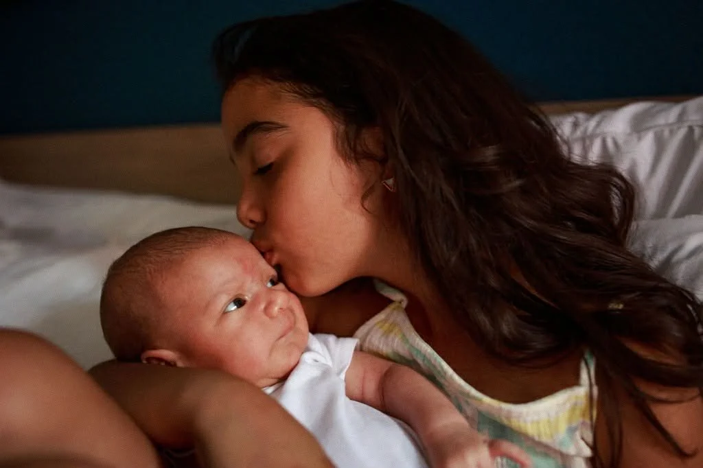 A young woman with long brown hair kissing a newborn baby on the forehead while lying on a bed.