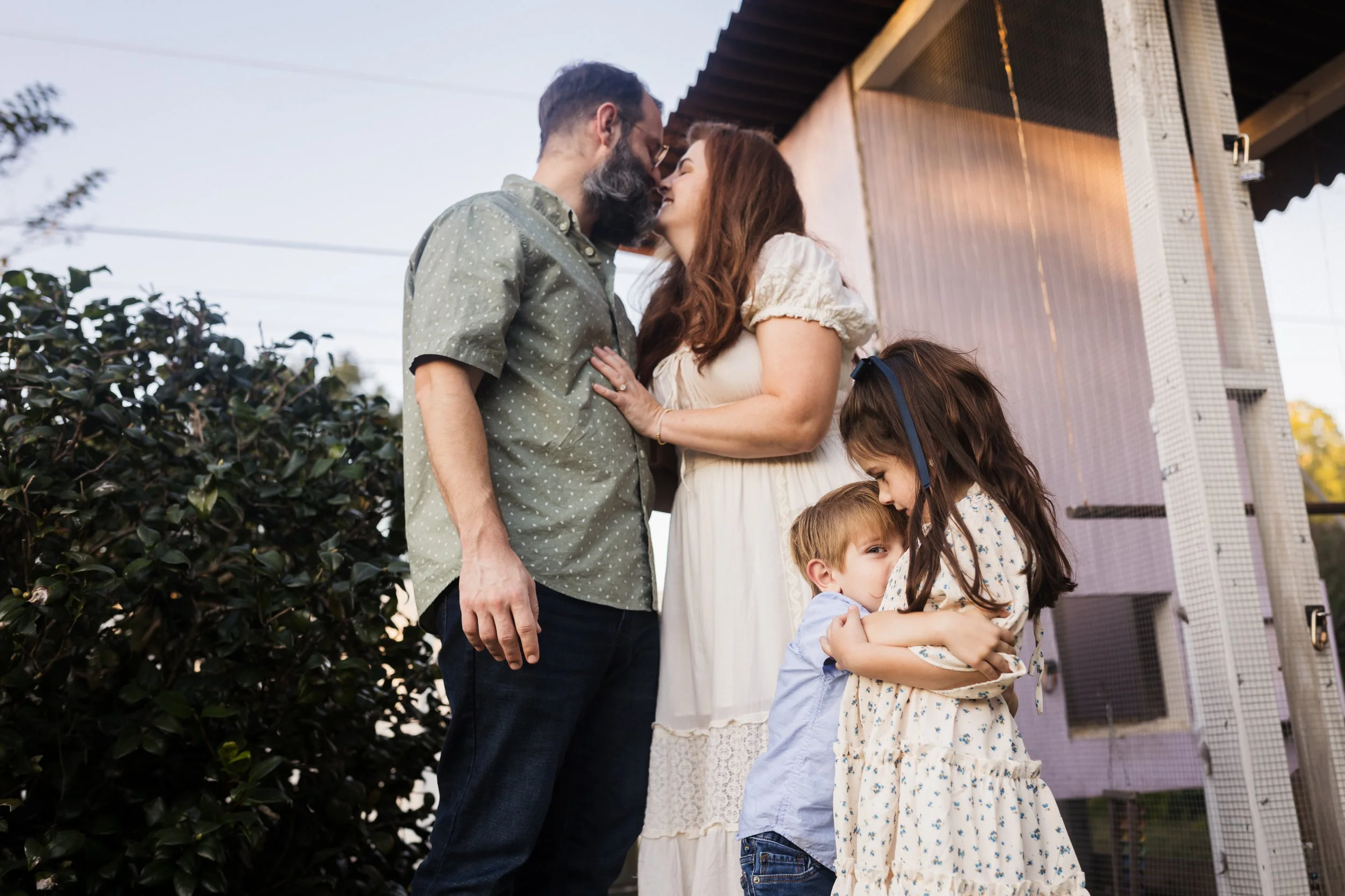 A family of four stands outside, with two adults and two children hugging and touching foreheads in an affectionate moment.