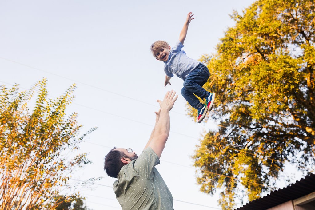 A man is tossing a smiling boy in the air outdoors during fall, with colorful autumn trees in the background.