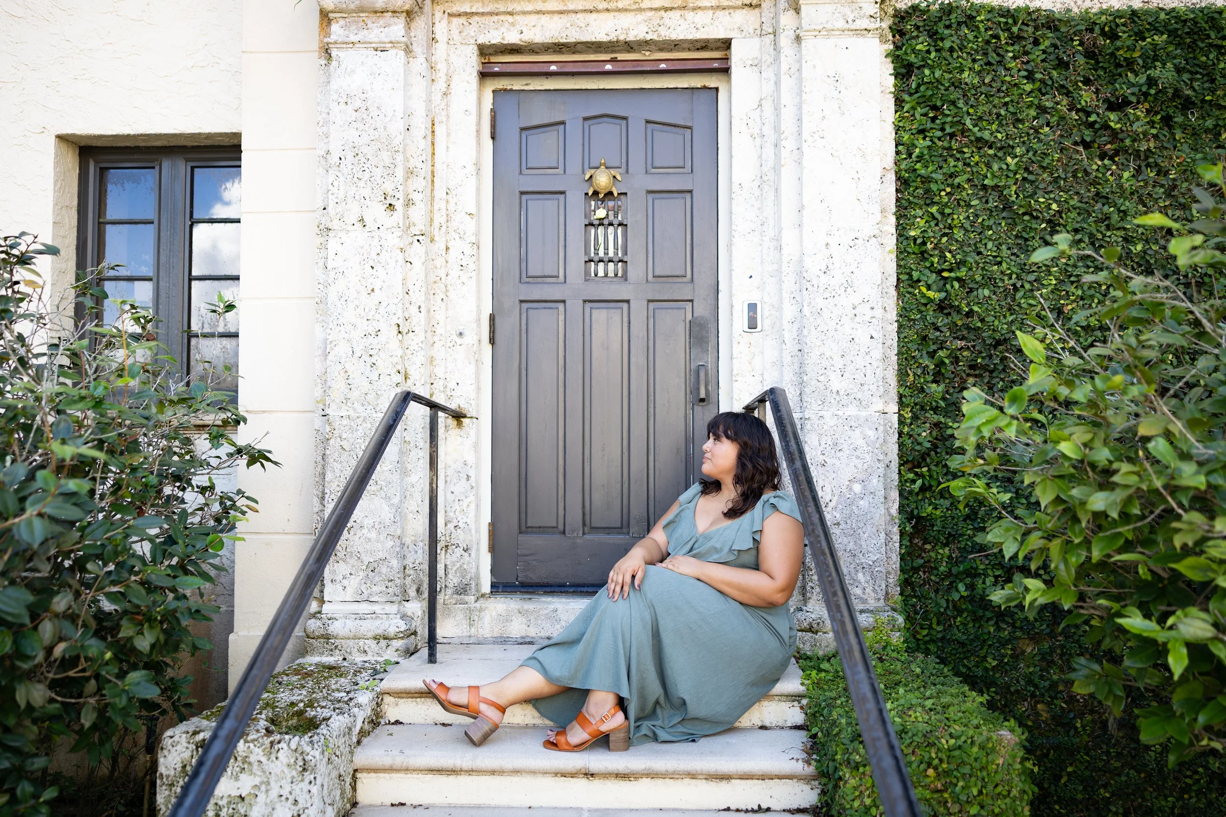 A woman in a green dress and sandals sitting on outdoor steps, leaning against a black railing in front of a dark gray door with a gold ornament, surrounded by white stone and green foliage.