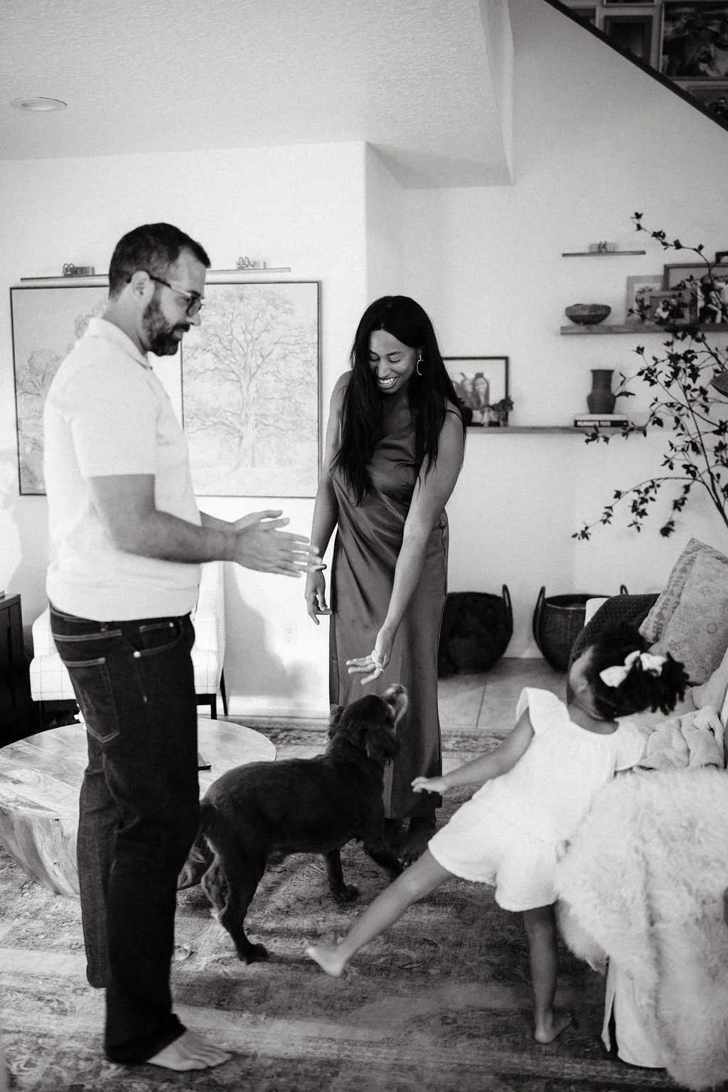 A family of four playing with a puppy in their living room. The man, woman, and two young girls are smiling and appear joyful. The room has wall art, shelves with decorative items, and a sofa with a floral pillow.