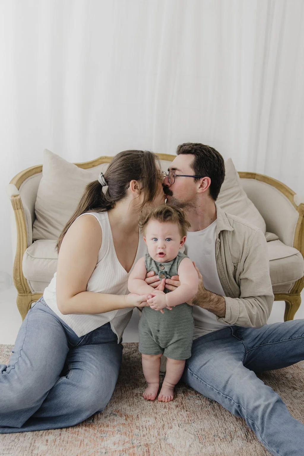 A family of three sitting on the floor in front of a sofa, with the parents kissing and the baby looking surprised.
