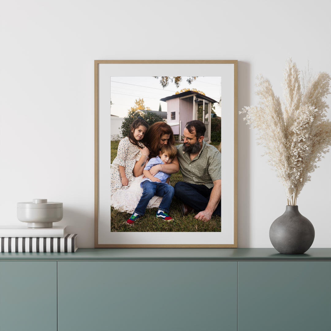 Family of four sitting on grass, smiling and hugging outdoors with a house and trees in the background.