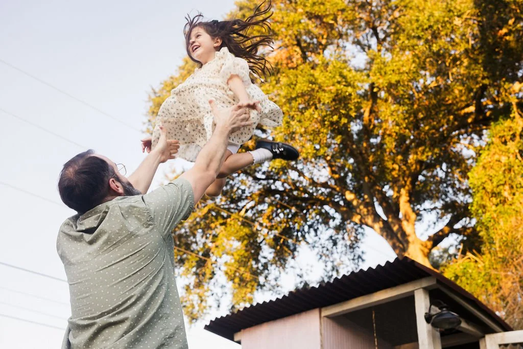 A girl in a white dress being tossed in the air by a man outdoors during fall, with trees in the background.