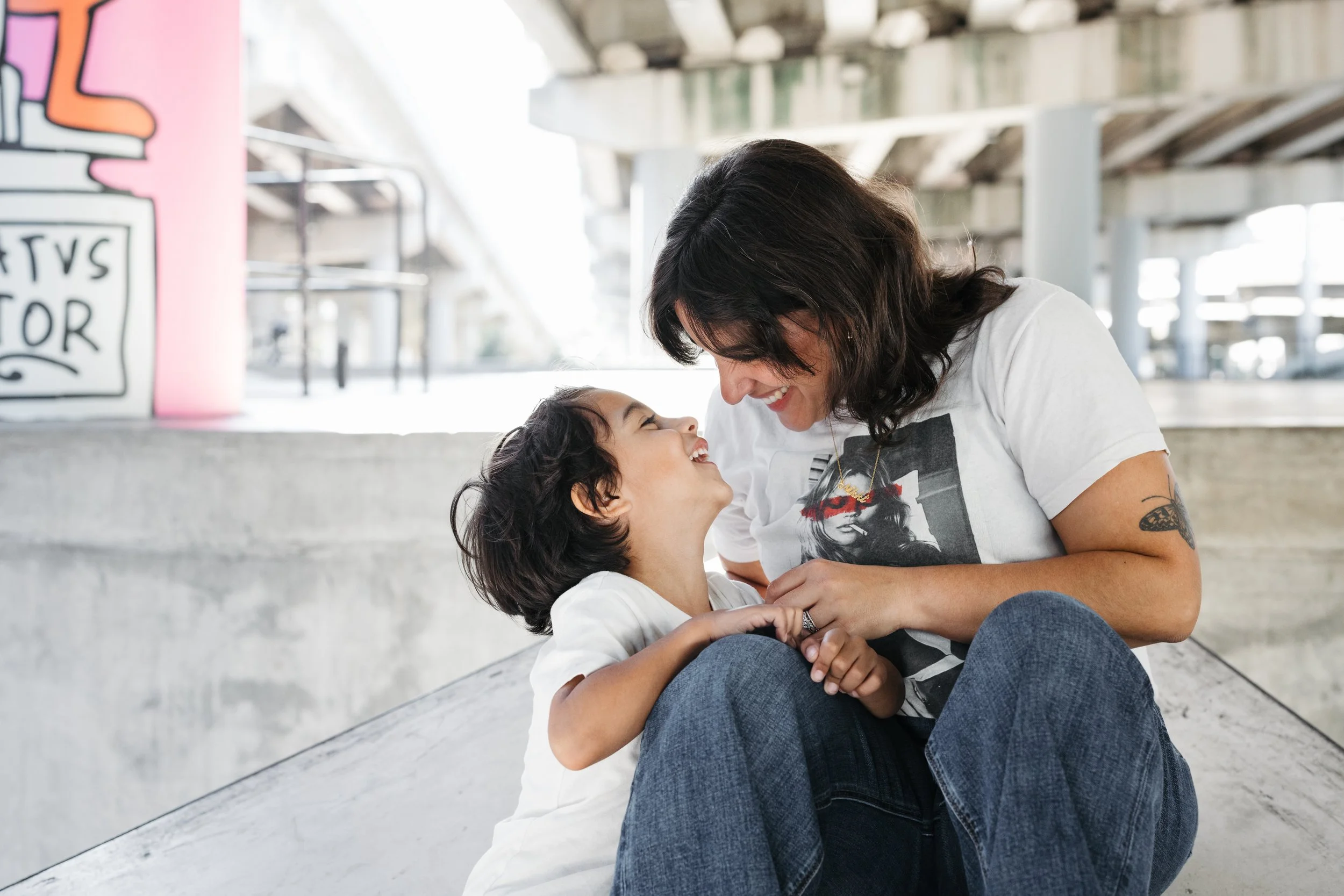 Mom and son sitting on a skatepark ramp, smiling and holding hands, under an overpass.