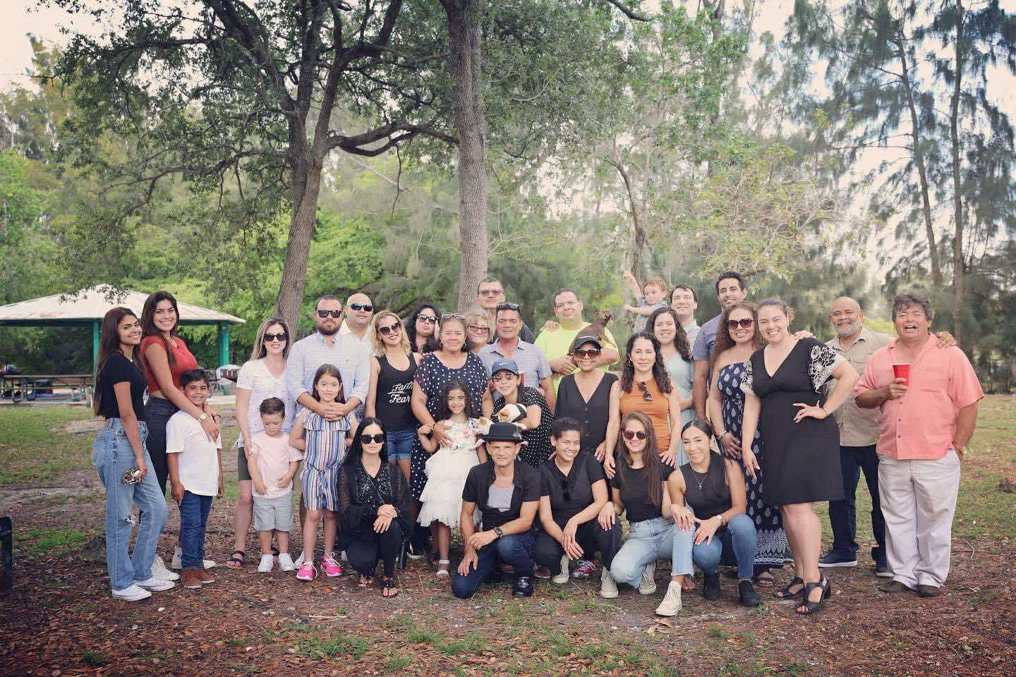 Large group of people gathered outdoors in a park with trees in the background, posing for a group photo.
