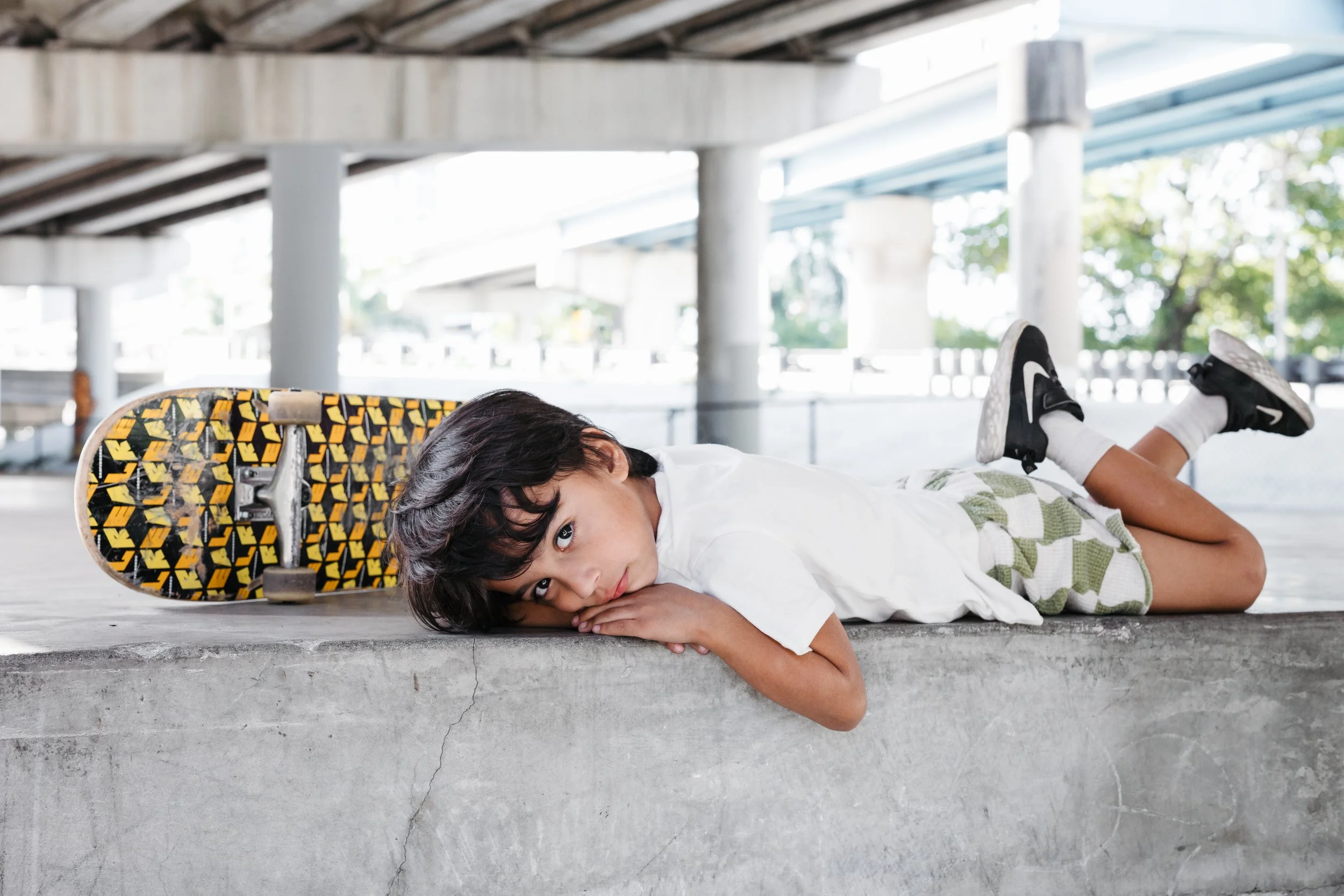 A young boy lying on his stomach on a concrete surface with a skateboard beside him under an overpass.