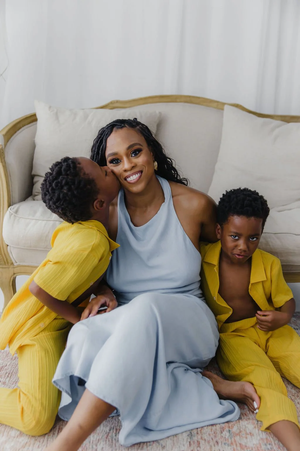 A woman with two young boys sitting on a carpeted floor in front of a beige couch with white pillows. One boy is giving the woman a kiss on the cheek, and the other boy has a slightly mischievous expression. The woman is smiling and looking at the camera.