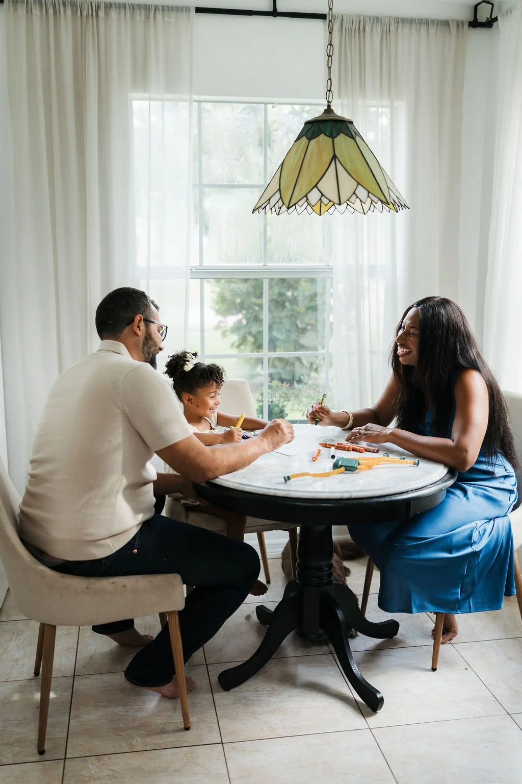 A family of three sitting at a round table near a large window, drawing and coloring with crayons, with a stained glass lampshade hanging above.