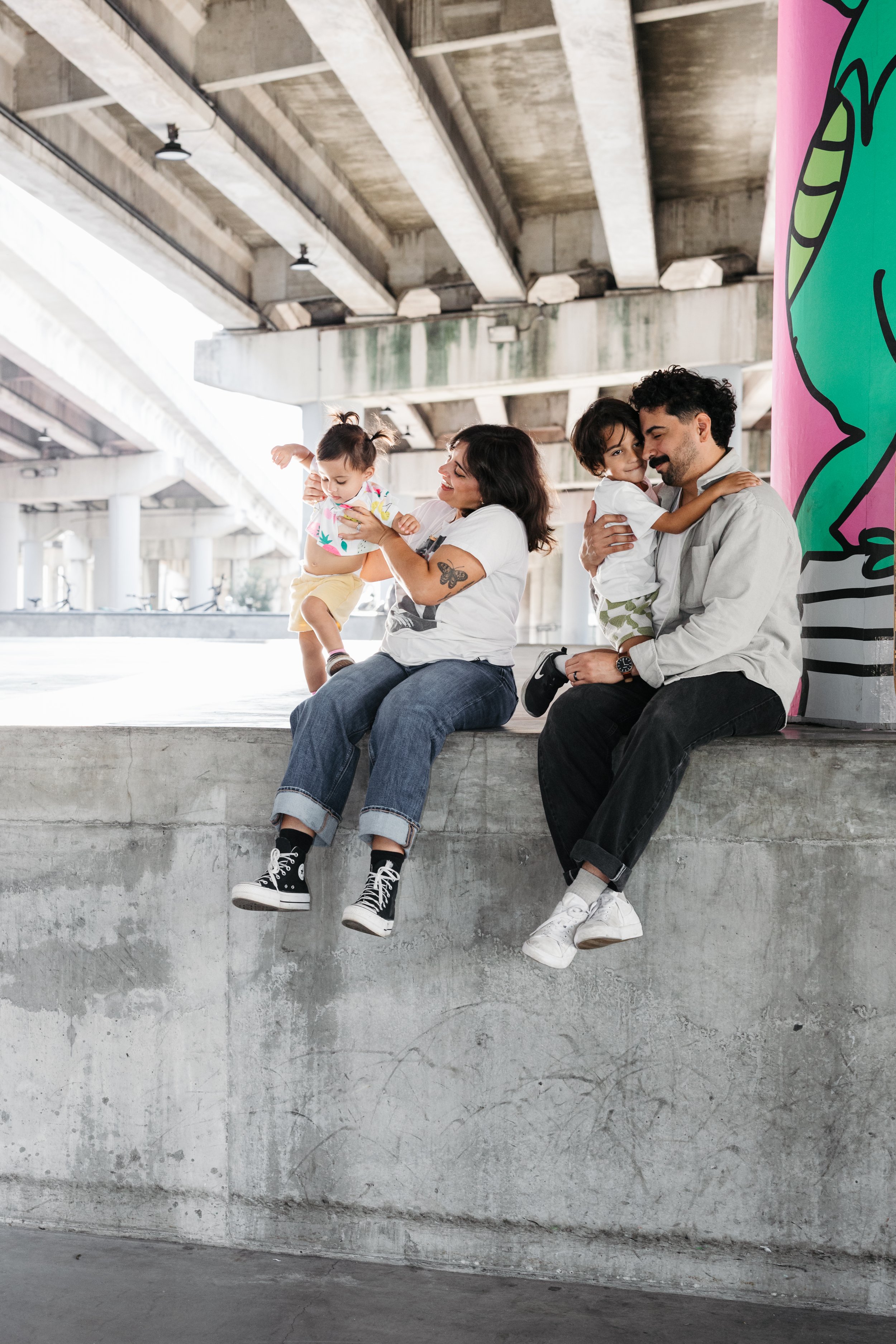 A family of four sitting on a concrete ledge under an overpass with colorful graffiti visible on one side. The mother holds a young girl while the father holds a young boy, all smiling and spending time together.
