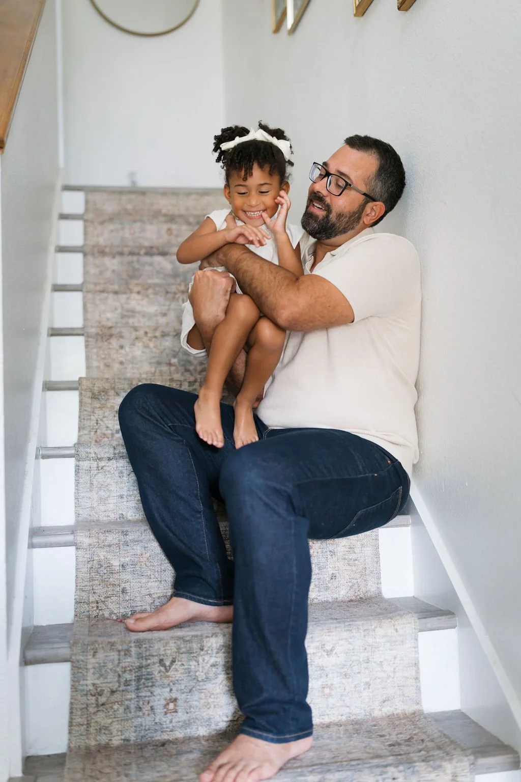 A man with glasses sitting on a staircase holding a young girl with a white bow in her hair, both smiling and playing together.