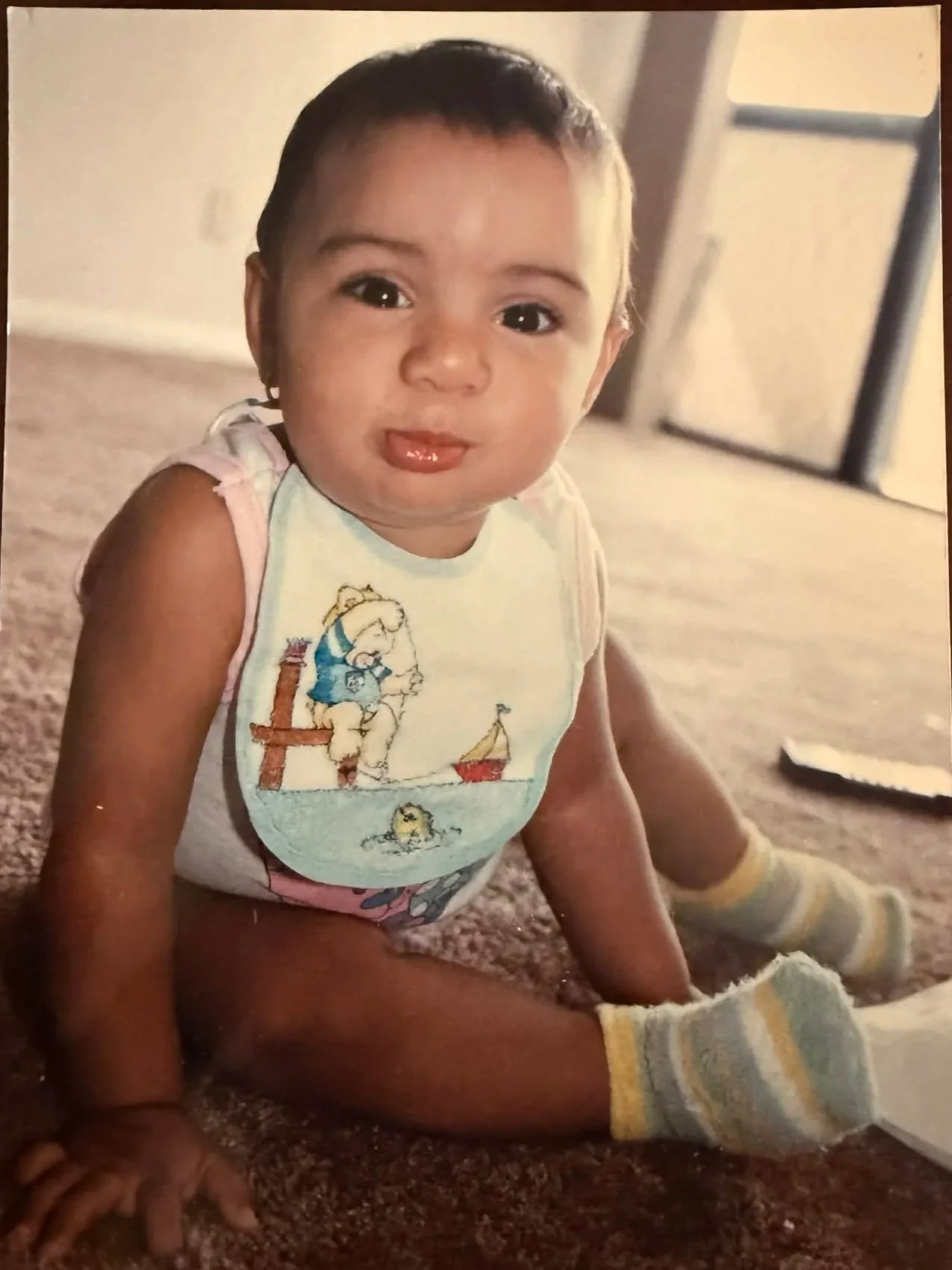 A young child with short dark hair, sitting on a carpeted floor, wearing a sleeveless shirt with a cartoon print, shorts, and striped socks, looking at the camera with a slight smile.