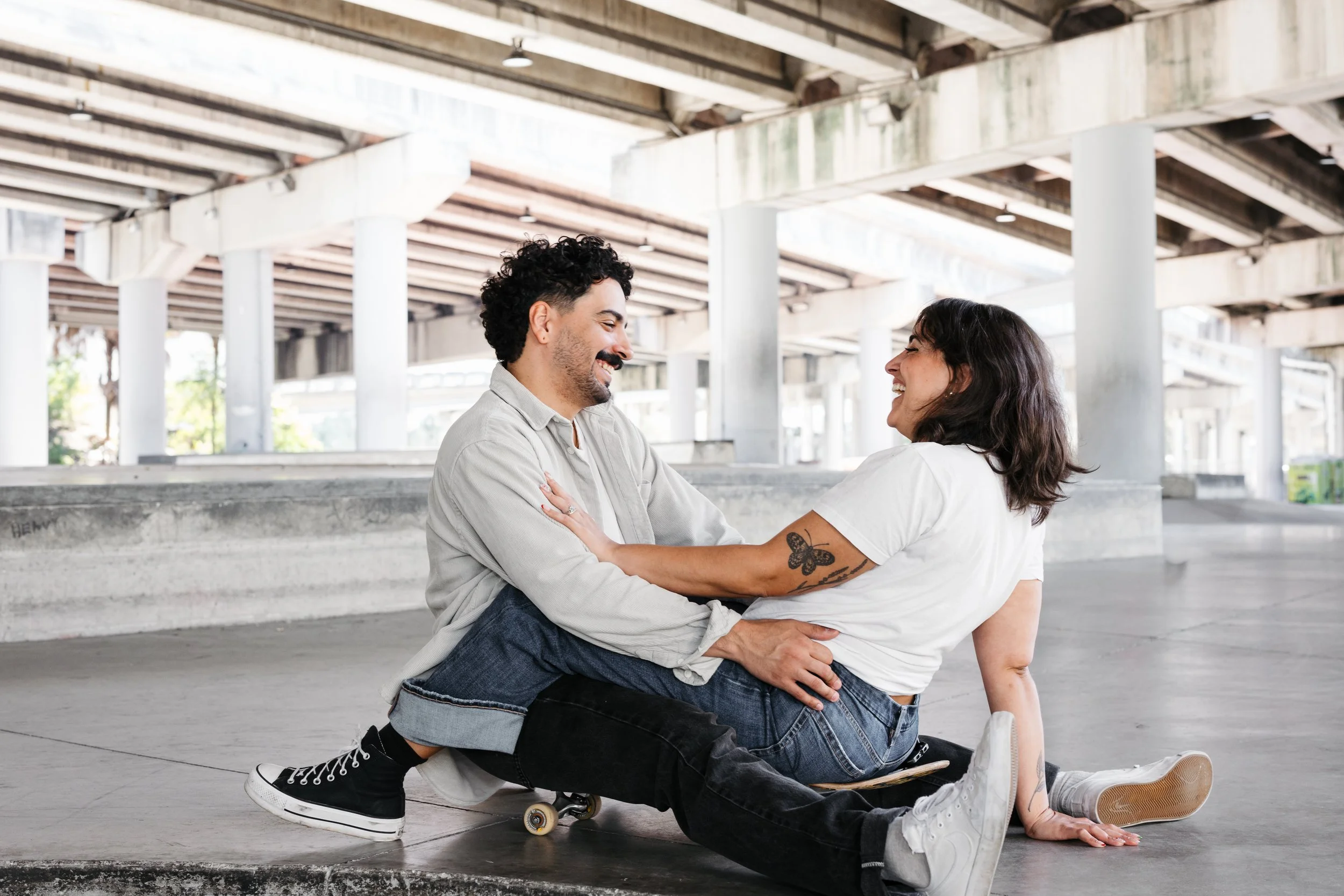 A man and a woman are laughing and smiling while sitting on skateboard under a bridge, with the woman sitting on the man's lap, in an urban setting.