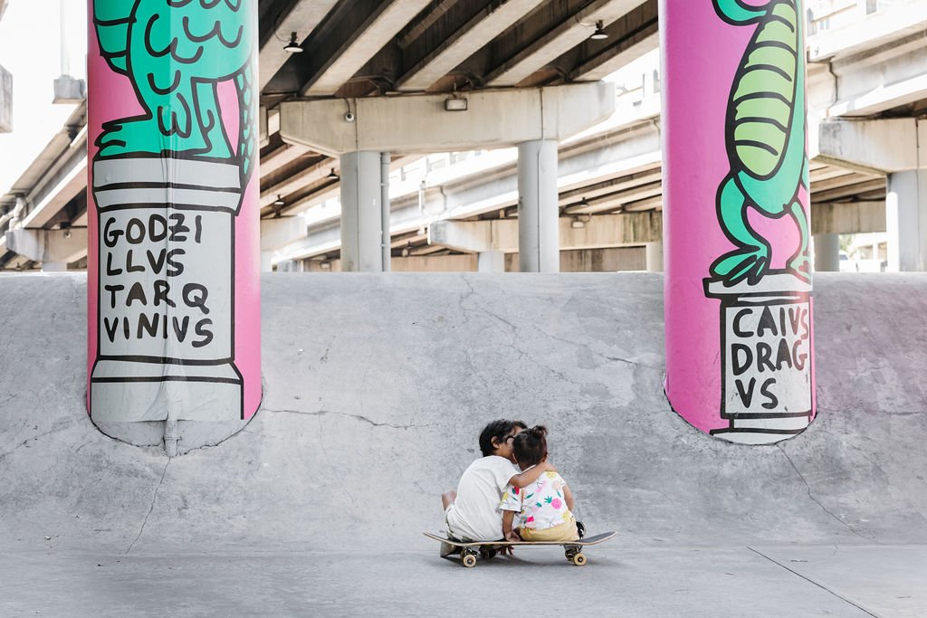 Two children sitting on a skateboard at a skate park, kissing, with large colorful murals of cartoon-style jars and a crocodile on the concrete wall behind them under an overpass.